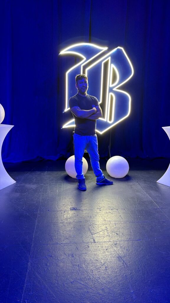 Líder de Fundación Equicentro posa con los brazos cruzados en el escenario de la final del Desafío Emprendedor de Banco de Chile. De fondo, un gran letrero de neón blanco con el logo del banco ('B') y una iluminación ambiental azul profesional. La imagen representa la visibilidad nacional y el profesionalismo de la organización desde Pencahue.