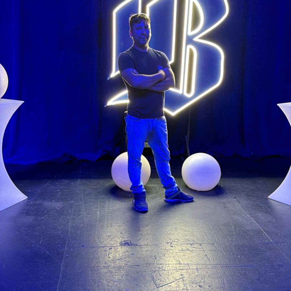 Líder de Fundación Equicentro posa con los brazos cruzados en el escenario de la final del Desafío Emprendedor de Banco de Chile. De fondo, un gran letrero de neón blanco con el logo del banco ('B') y una iluminación ambiental azul profesional. La imagen representa la visibilidad nacional y el profesionalismo de la organización desde Pencahue.